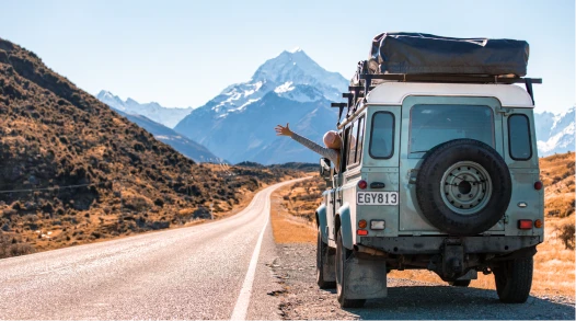 A person waving from the window of a Land Rover with New Zealand mountains in the background.